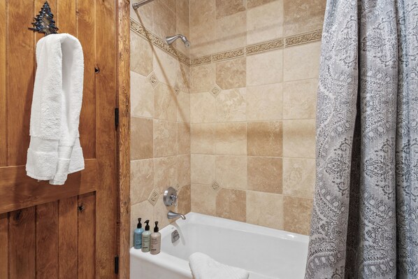 Un baño con puerta de madera, una toalla blanca colgada en la puerta, una bañera con cortina de ducha y artículos de aseo en el borde de la bañera.