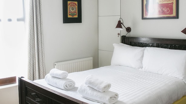 Een net opgemaakt bed met witte lakens en opgerolde handdoeken, een donker houten hoofdbord, een rode hanglamp, een ingelijst poster aan de muur en een raam met gordijnen.