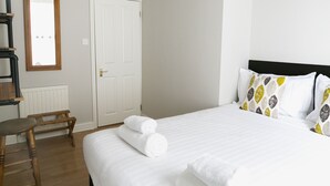 Een hotelkamer met een bed, witte lakens, een houten bankje, een witte deur en een spiegel aan de muur.