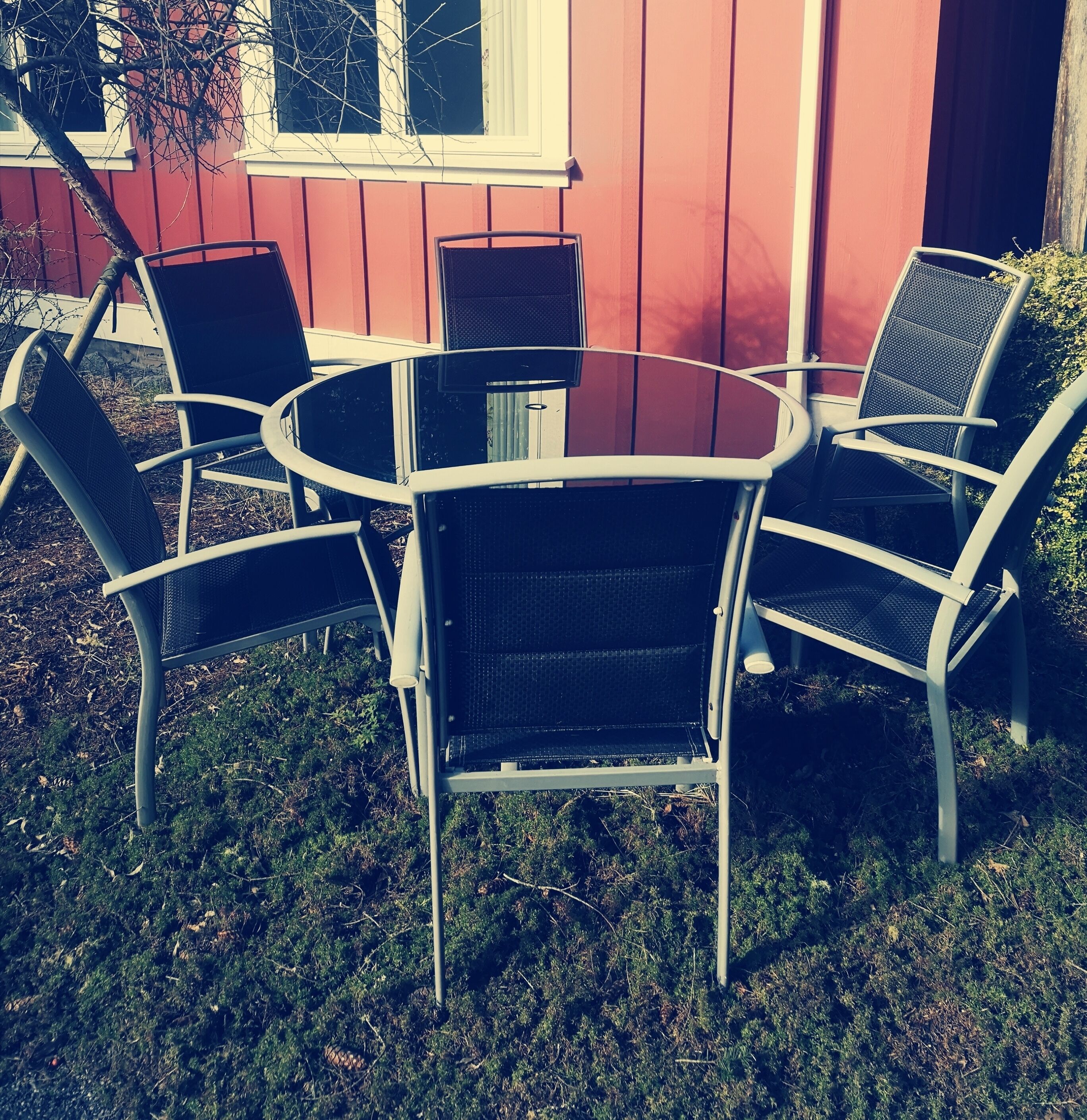 Une table ronde en métal, entourée de quatre chaises, est placée à l’extérieur, devant un bâtiment rouge doté d’une fenêtre.