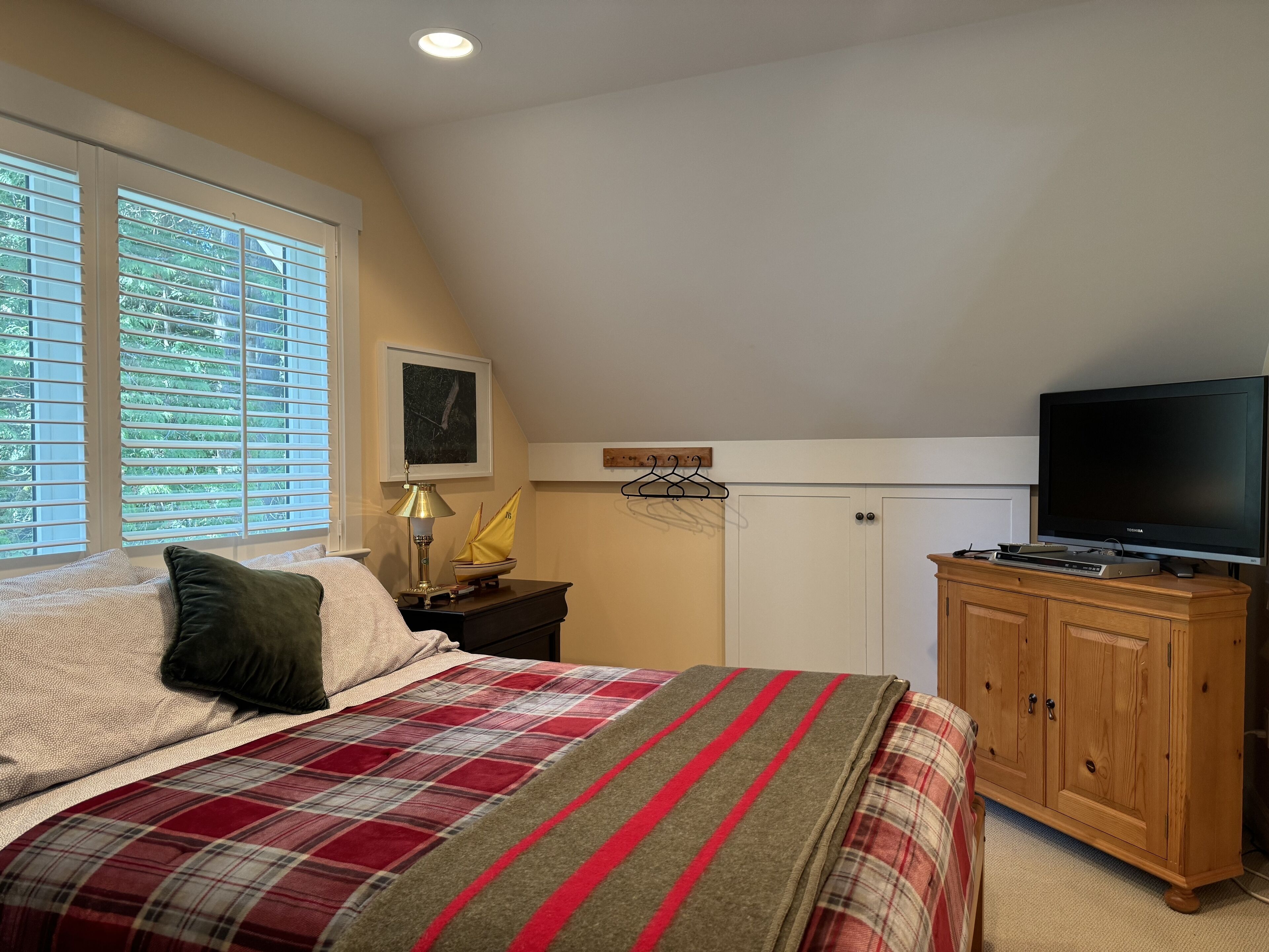 Un dormitorio con cama, televisor sobre un mueble de madera, ventana con persianas y un cuadro en la pared.
