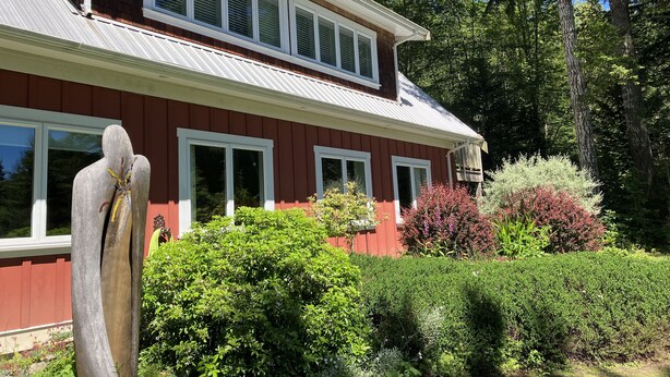 Une maison à deux étages, avec une façade rouge et des éléments décoratifs blancs, entourée d’une végétation luxuriante et de grands arbres.
