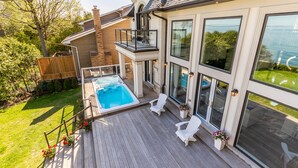 Une maison moderne dotée d’une grande porte vitrée, d’un balcon avec un jacuzzi et d’une terrasse en bois avec des chaises blanches.