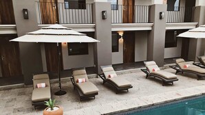 Un espace aménagé au bord de la piscine, avec des chaises longues et des parasols, un pot de fleurs et un bâtiment doté de balcons.