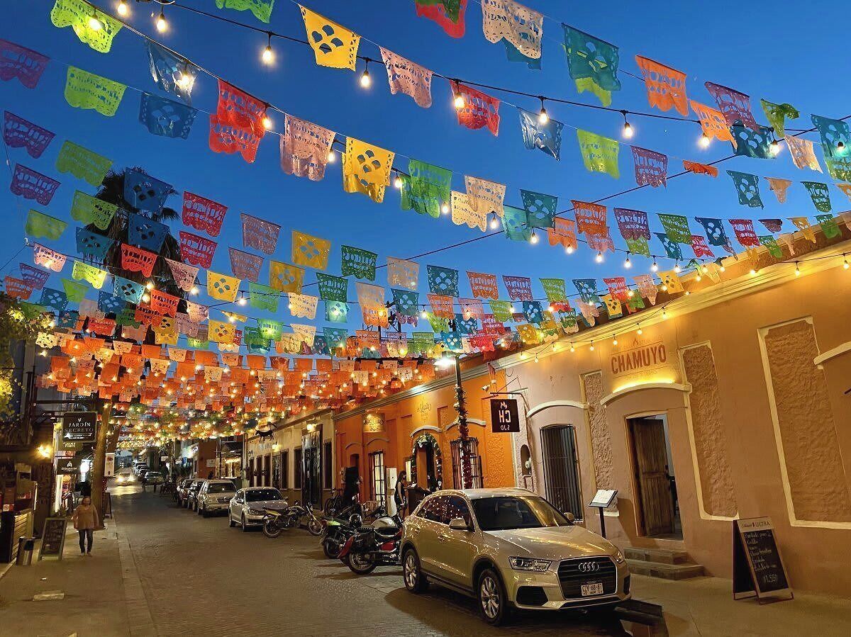 Calle bordeada por vehículos estacionados y edificios, decorada con banderines de colores.