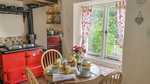 Una cocina con estufa roja, sillas de madera, una mesa de comedor con cosas para el desayuno y una ventana con cortinas florales.