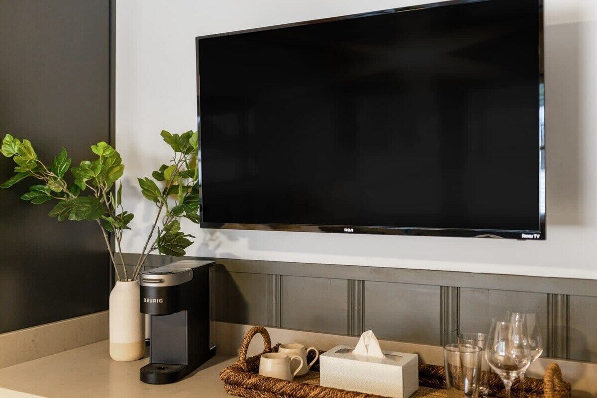 A modern living room with a flat-screen TV mounted on the wall, a Keurig coffee maker, a vase with green plants, and a tray with a tissue box, mugs, and glasses.