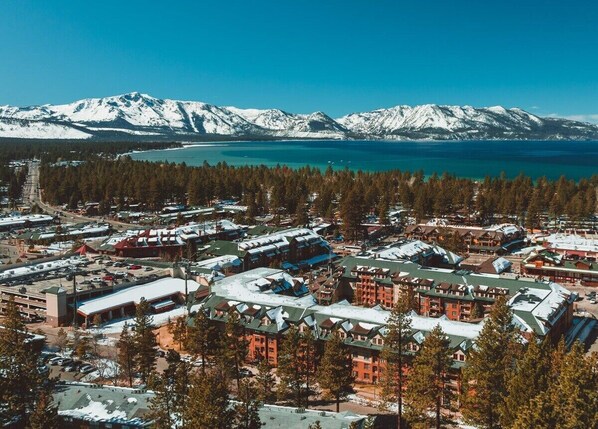 A resort area with multiple buildings, a parking lot, and a backdrop of snow-capped mountains and a lake.