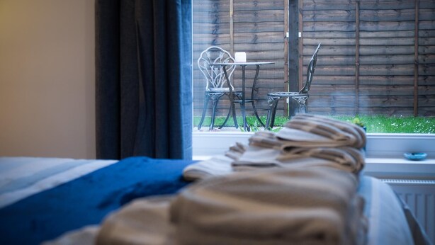Una cama con un edredón azul y otra manta doblada encima. Se ve una ventana con vistas a una valla de madera y dos sillas afuera.