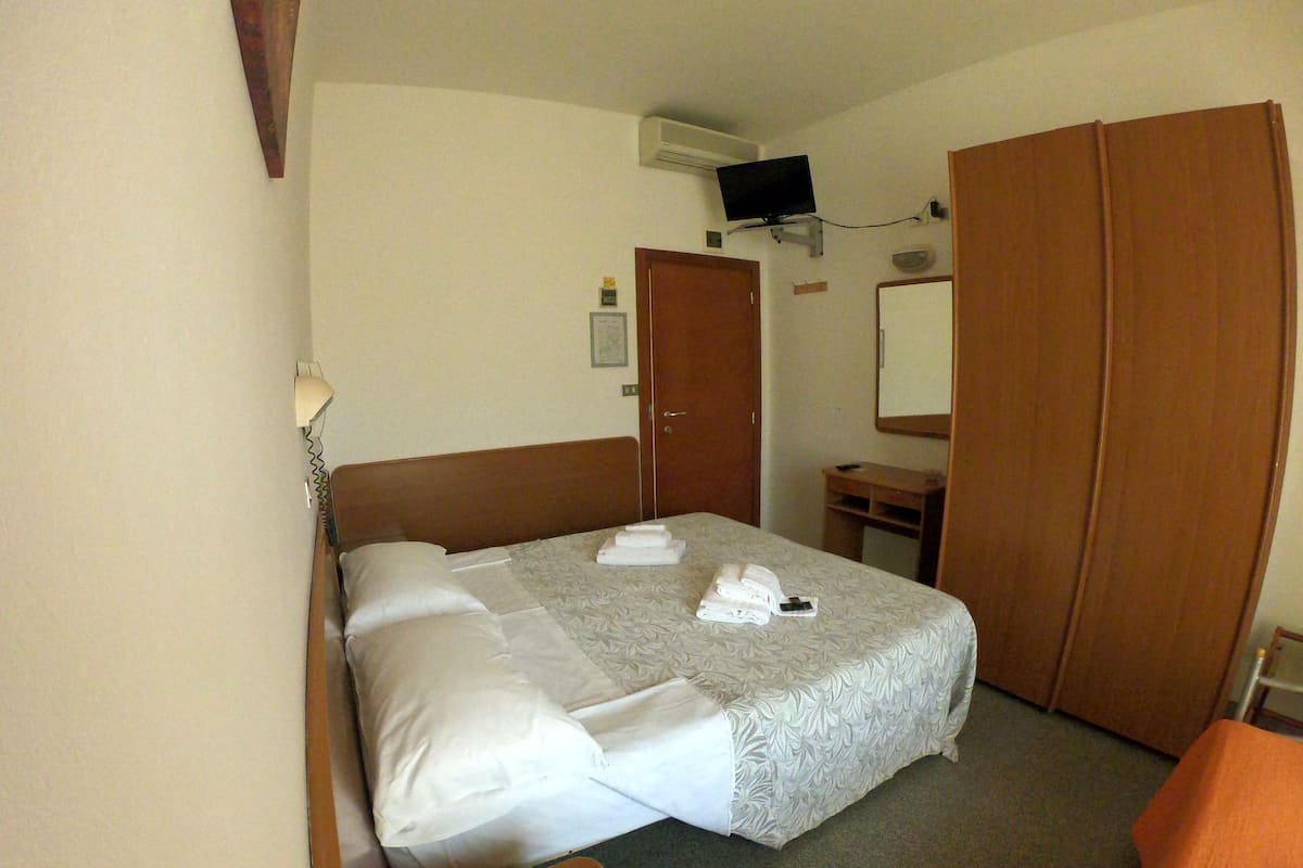 A hotel room with a bed, a wooden wardrobe, a small desk, a mirror, and a television mounted on the wall.