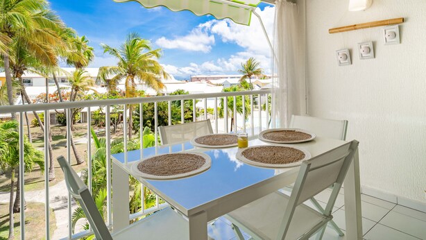 Un balcone con un tavolo apparecchiato per quattro, affaccia su un giardino con palme e un cielo azzurro intenso.