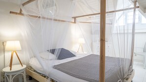 Un letto a baldacchino con tende, una coperta e cuscini bianchi, due abat-jour e un tavolino bianco con una lampada.