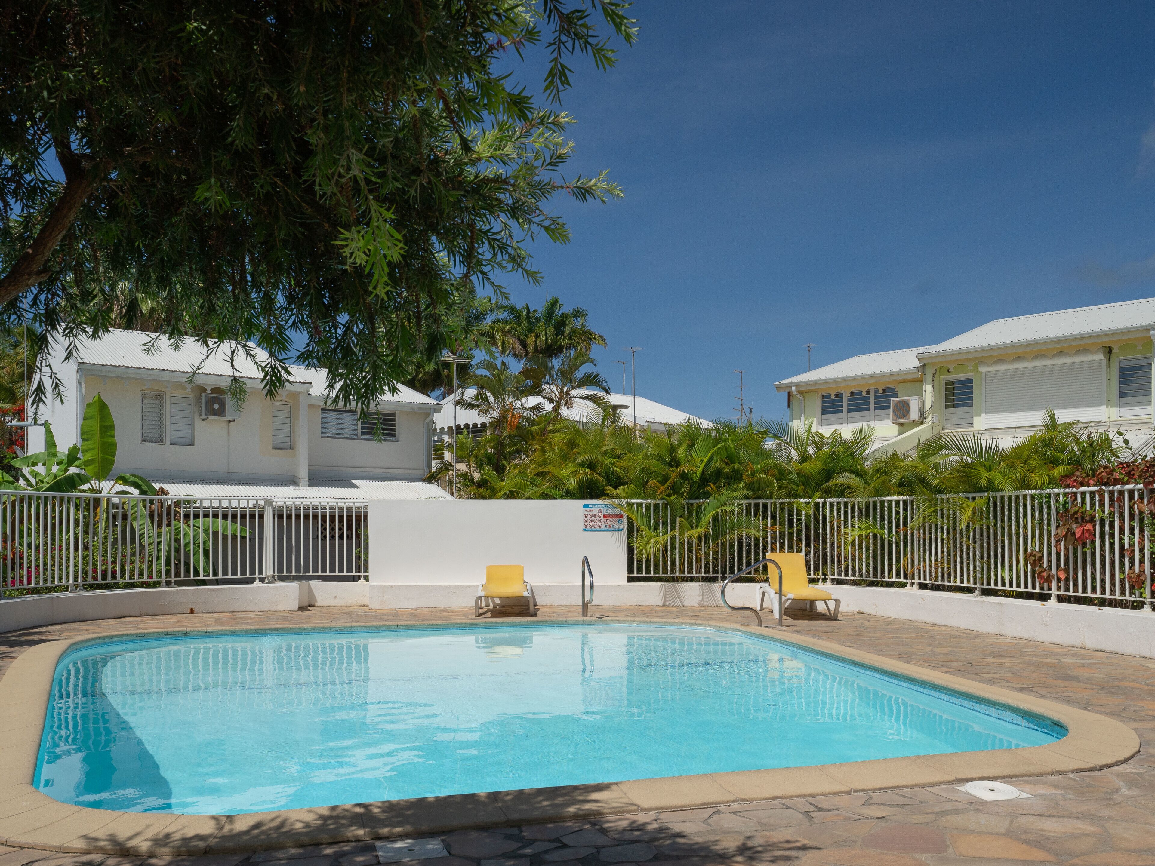 A swimming pool with a tiled deck, surrounded by a white fence, and a tree providing shade.