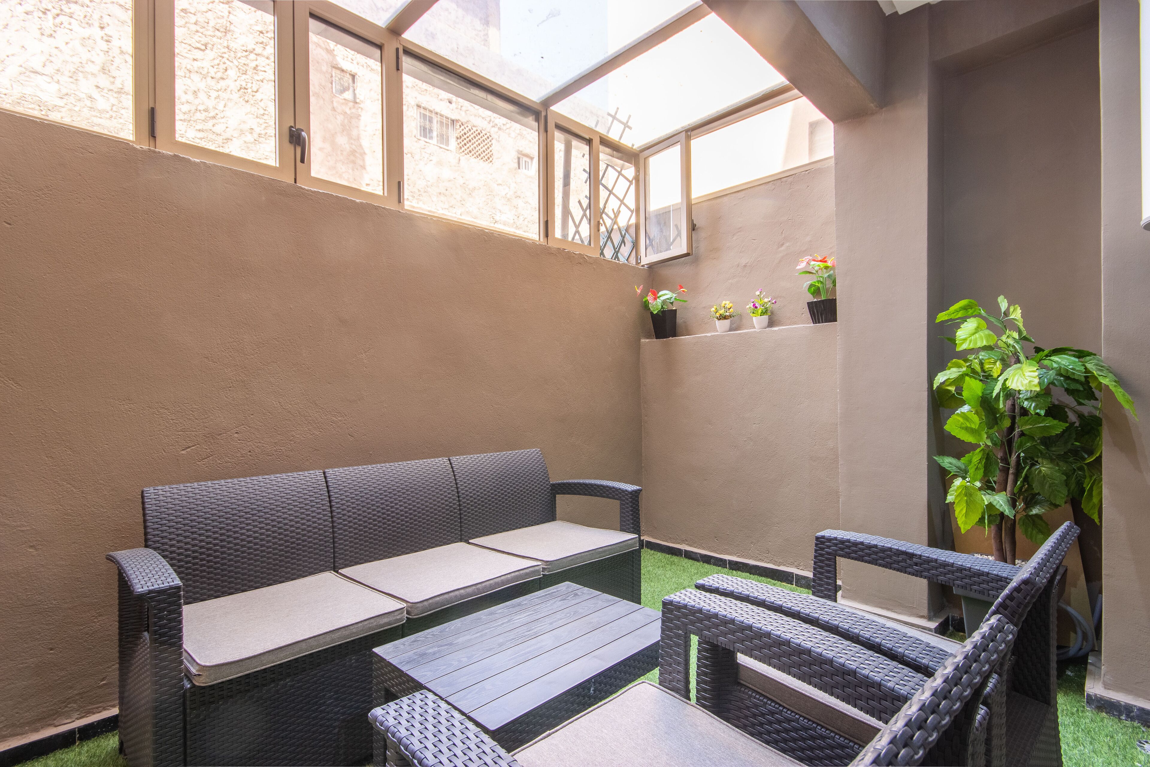 A patio area with a wicker sofa, a small table, and chairs. There are potted plants on a shelf and a large plant to the side.