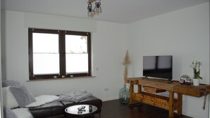 A modern living room with a leather sofa, a small round table, a wooden TV stand, and a large window with blinds. - Vacation home in Wehr Eifel, near Laacher See and Nürburgring with large garden (Wehr)