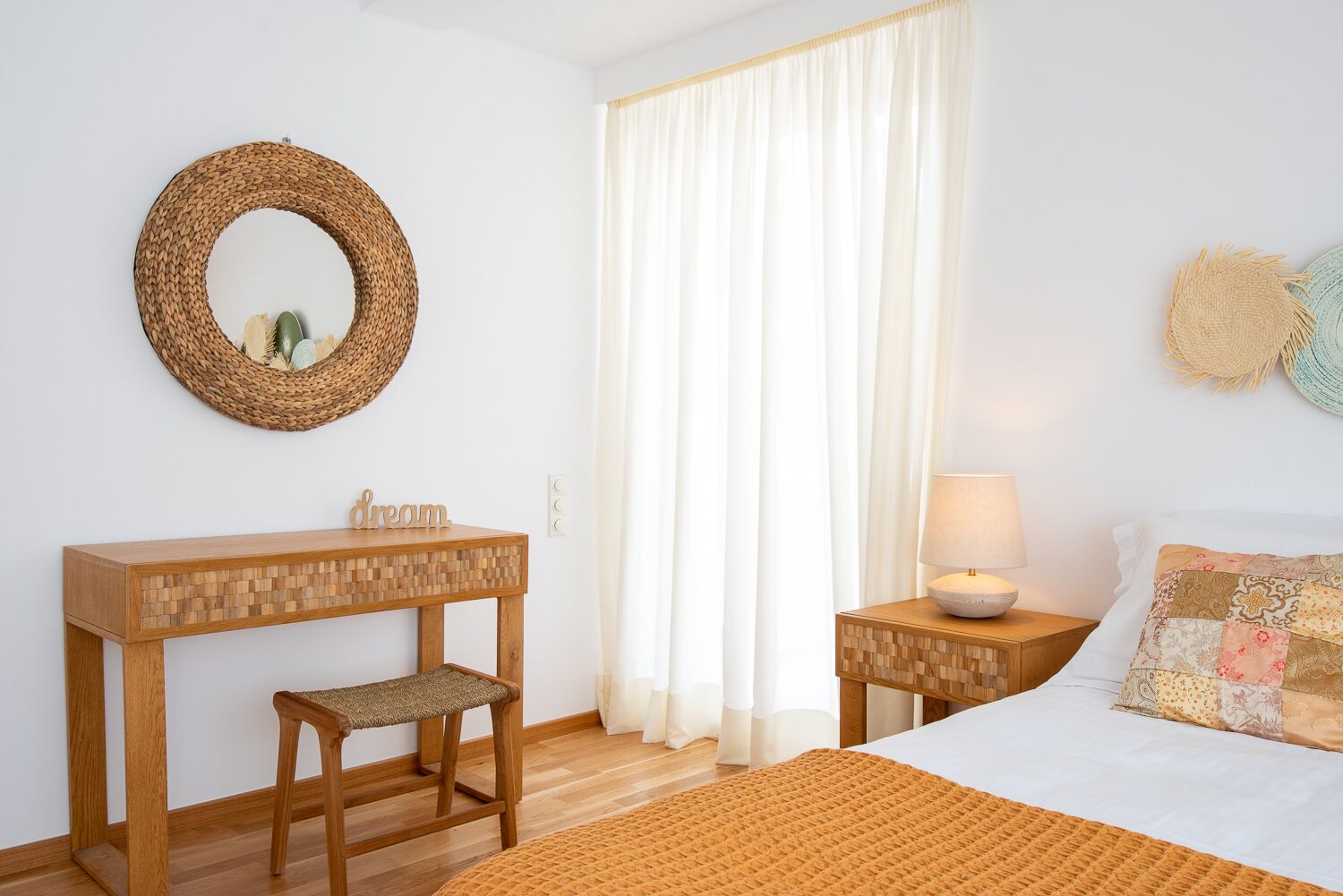 A bedroom with a wooden desk, a woven mirror, a bedside table with a lamp, a bed with a patterned pillow, and a window with white curtains.
