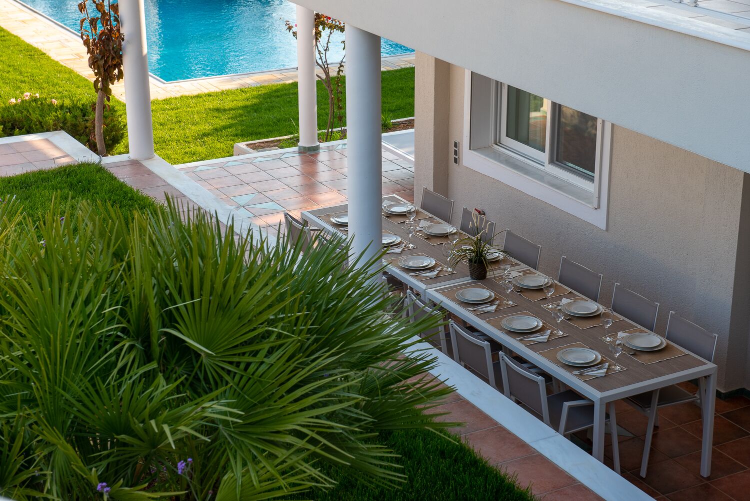 Un espace de restauration en plein air avec vue sur la piscine, des colonnes blanches et une longue table dressée pour le repas.