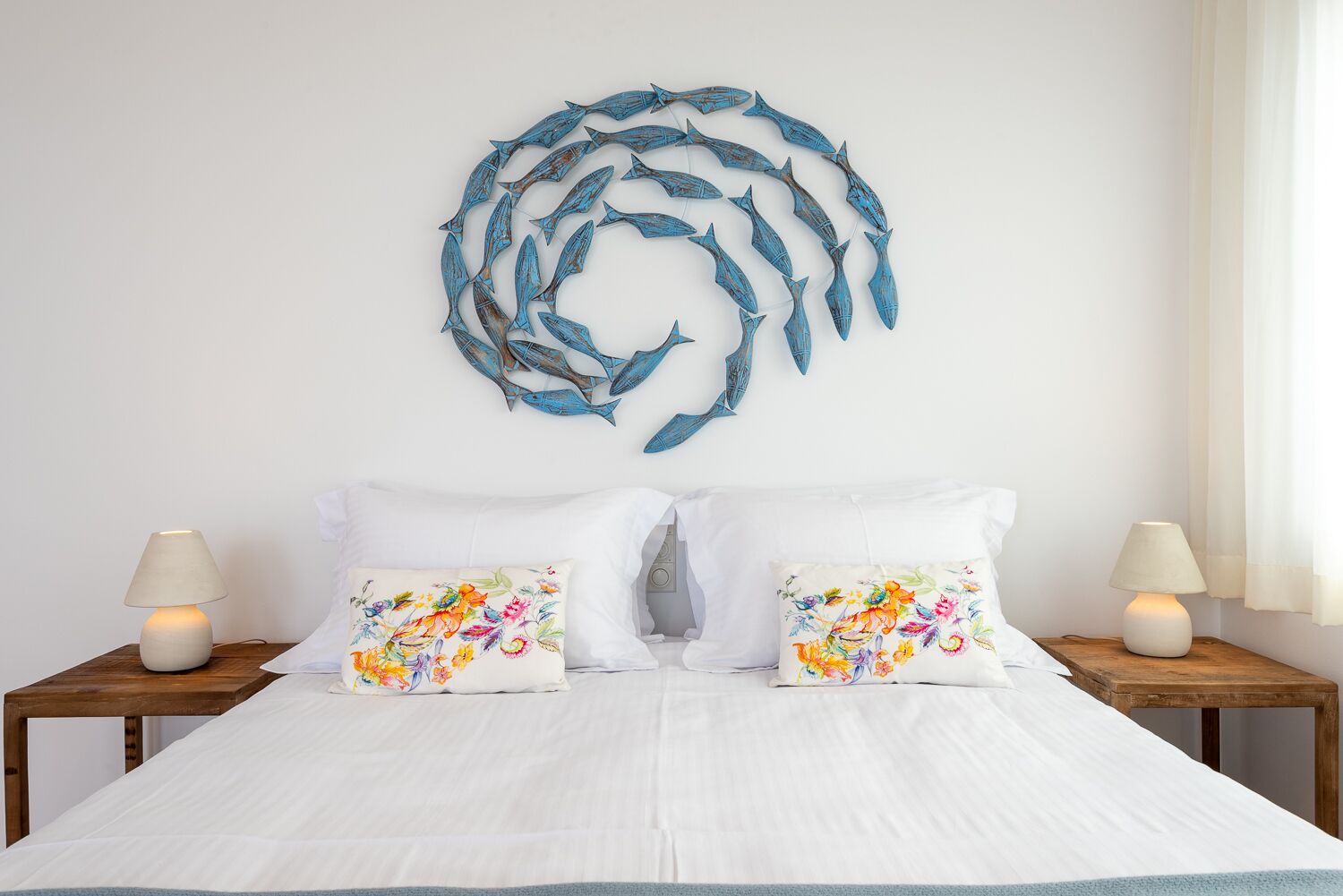 A bed with white linens, two matching pillows with floral designs, and a wall art of blue fish.