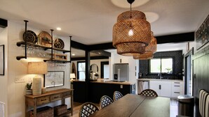 A dining area with a wooden table, chairs, and a hanging light fixture. There is a kitchen area with white cabinets and a black countertop.