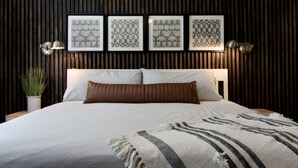 A neatly made bed with a striped throw, a potted plant, and framed geometric art on the wall.