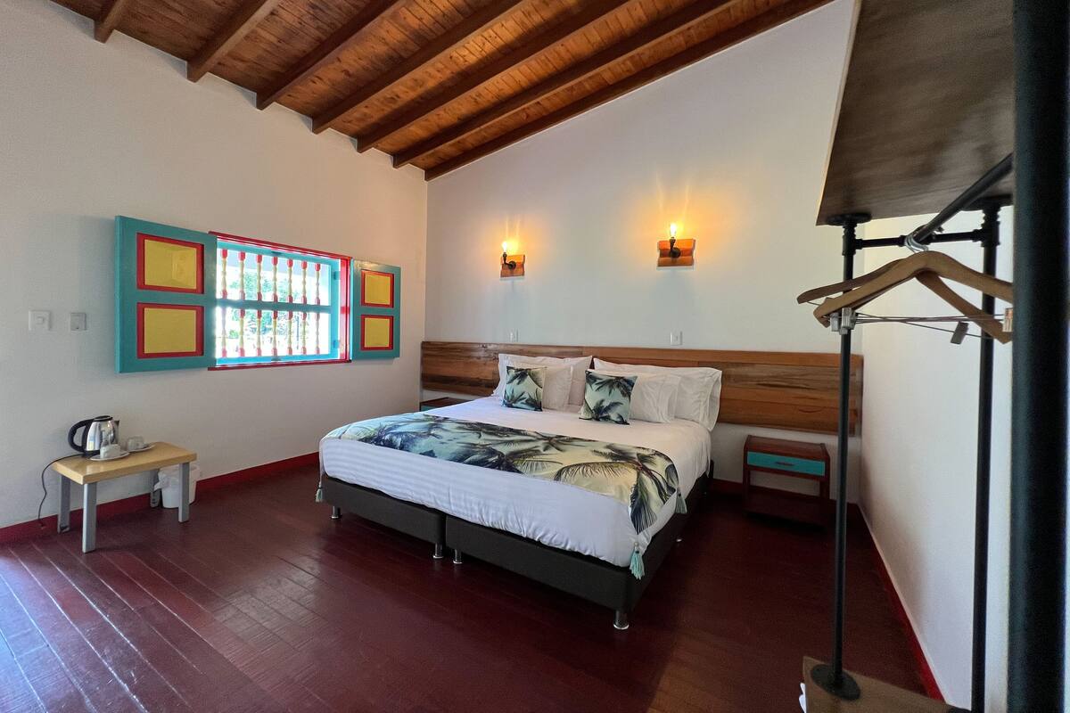 A bedroom with a wooden ceiling, a bed with a white and green floral bedspread, two wall-mounted lights, a small table with a kettle, and a colorful artwork on the wall.