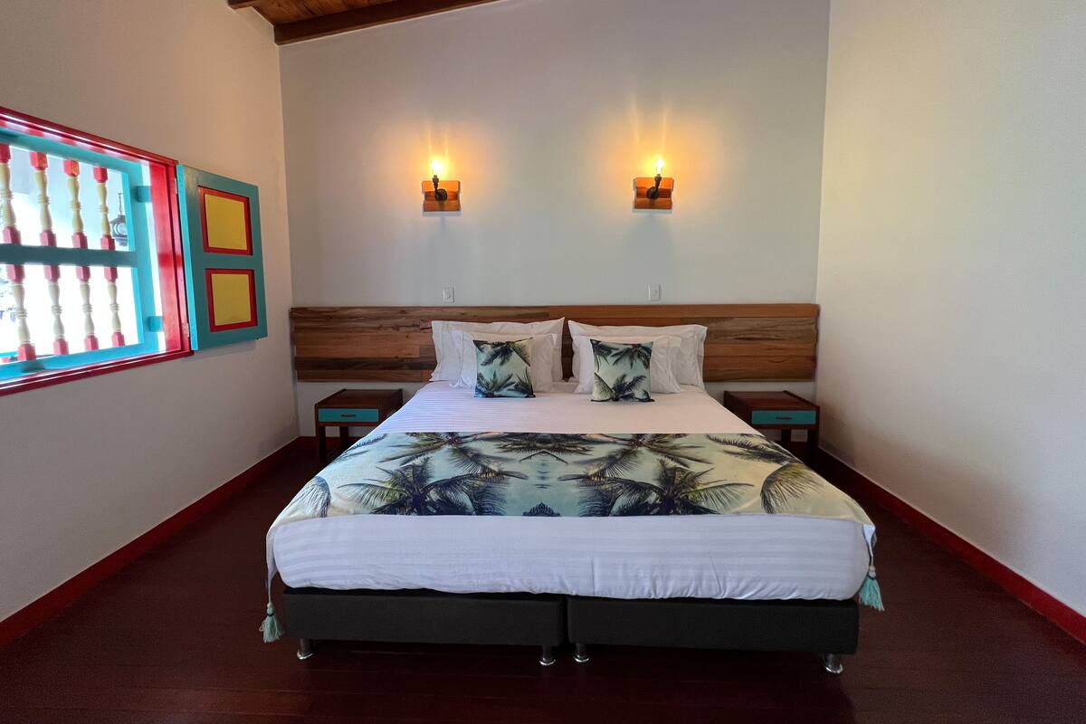 A bedroom with a wooden headboard, two bedside lamps, a bed with a tropical print bedspread, and a window with a colorful view.