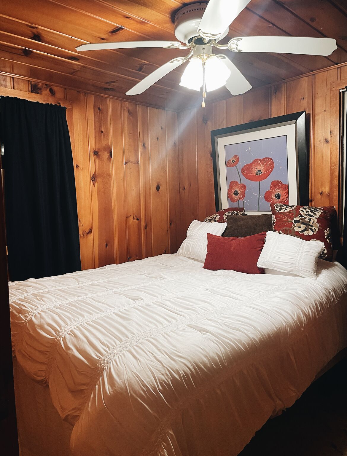 A wooden-paneled bedroom with a bed, a ceiling fan, and a framed artwork of red flowers.