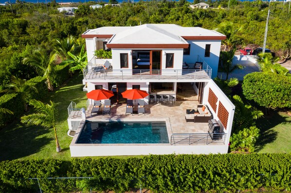 A two-story house with a swimming pool, outdoor seating, and a red car parked nearby.