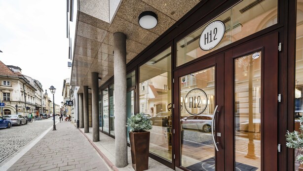 A hotel entrance with a large glass door, a sign reading "H12", and a potted plant on the sidewalk.