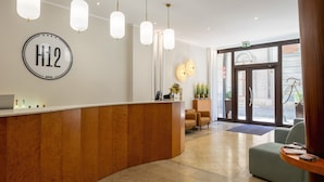 Zona de recepción de hotel con un mostrador de madera, un letrero grande H12 y un área de descanso con dos sillones.