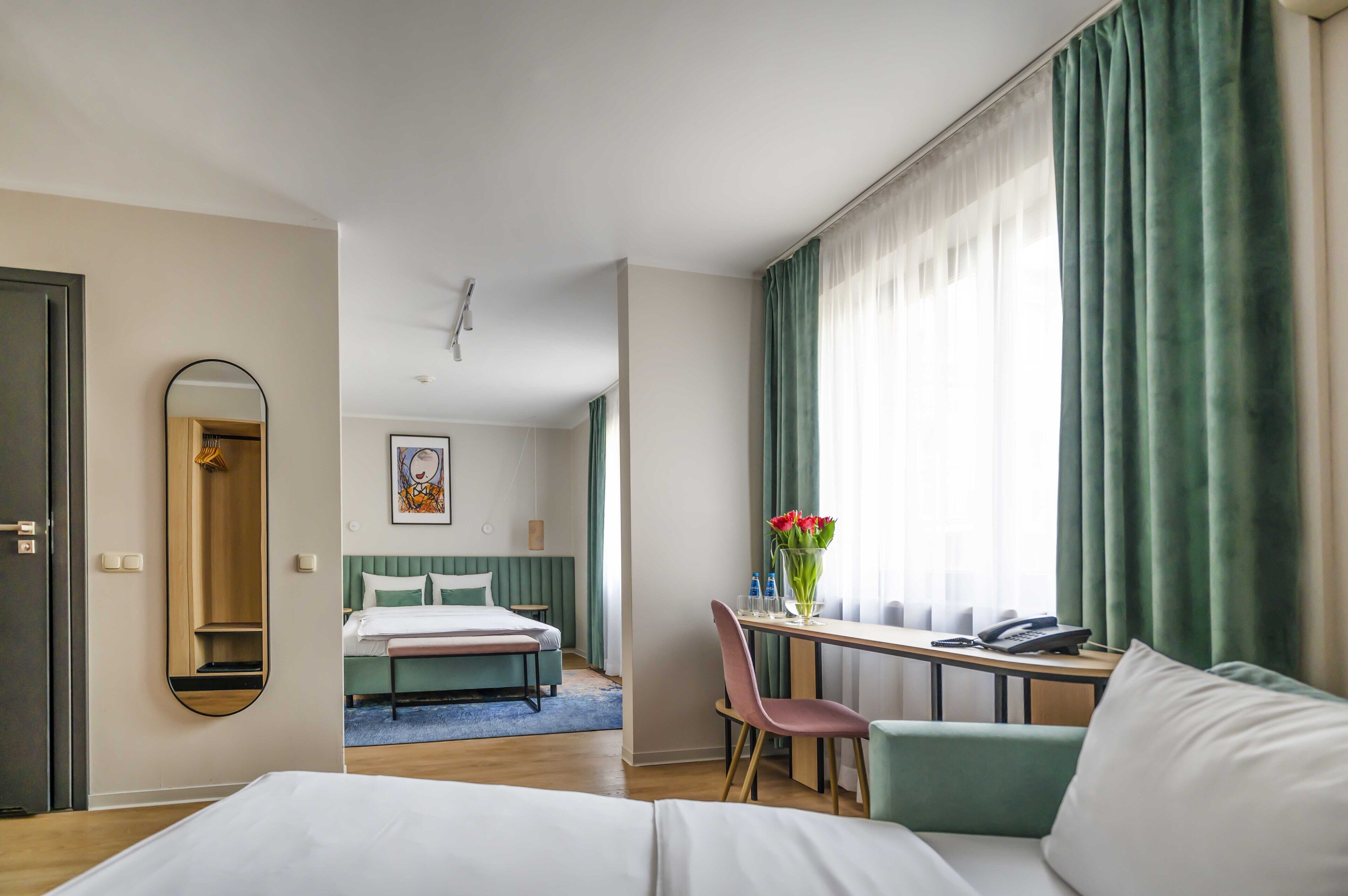 Une chambre d’hôtel comprenant un lit, un bureau avec une chaise, un miroir, une fenêtre avec des rideaux et un tableau au mur.
