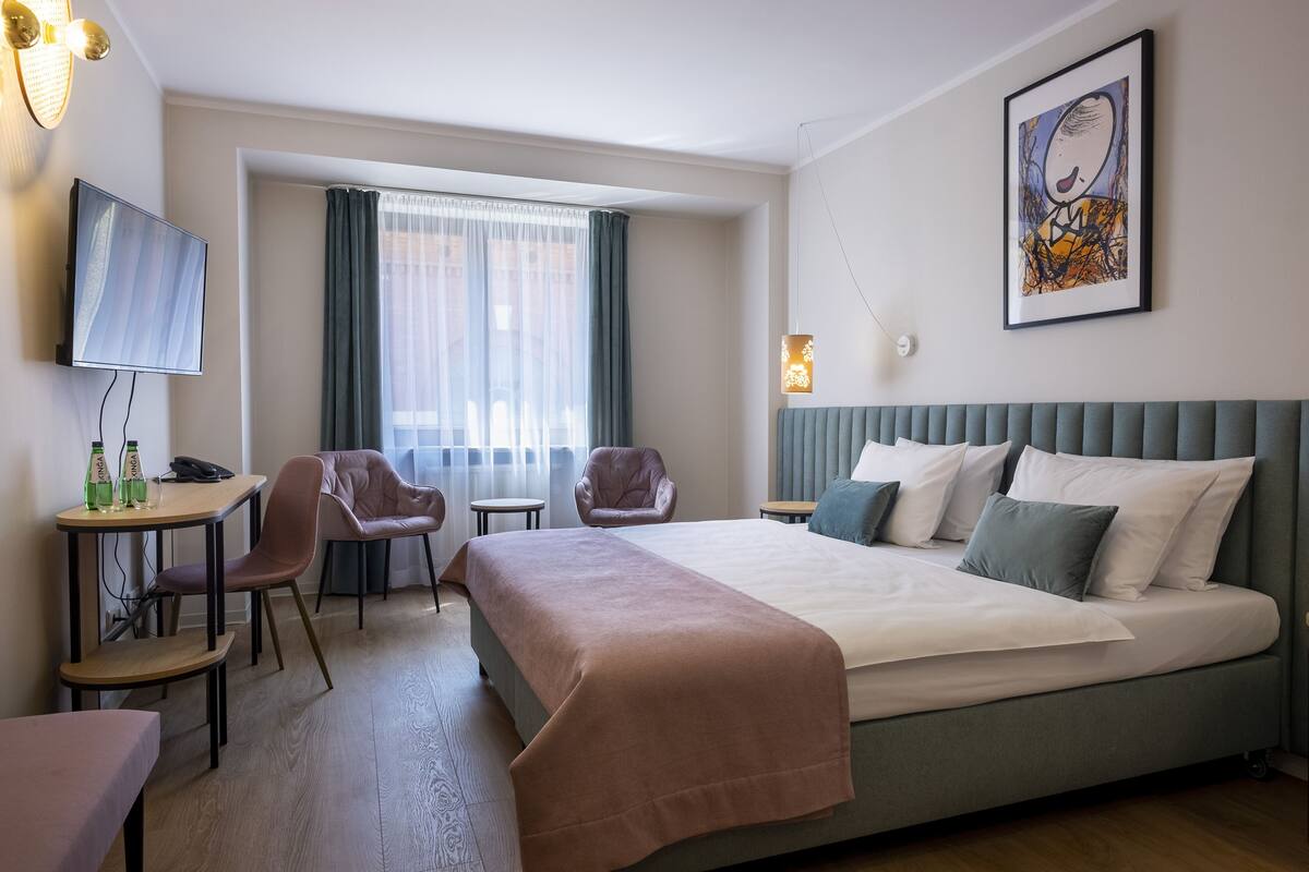 Una habitación de hotel con cama, escritorio y sillas, televisor y ventana con cortinas.