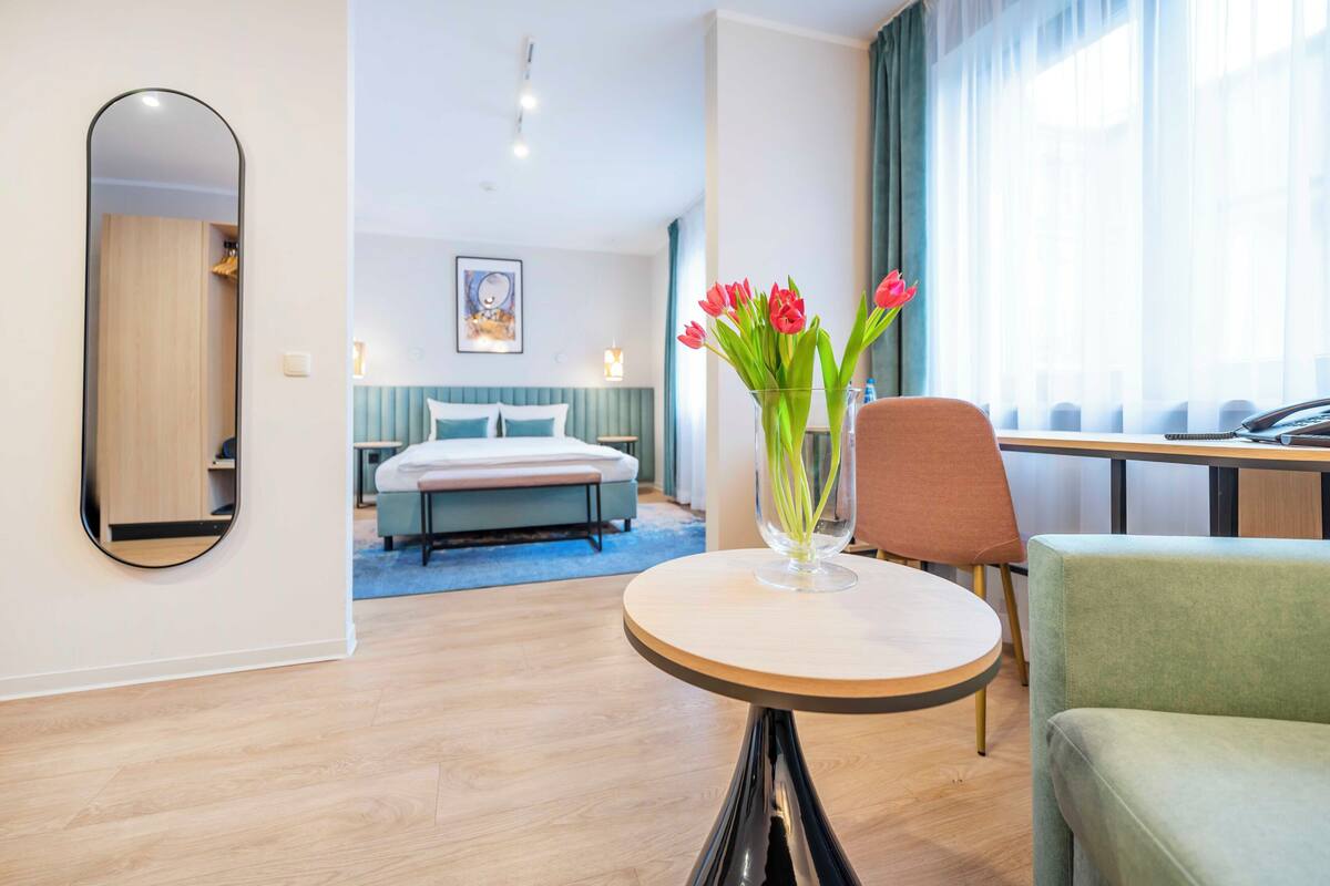 Una habitación de hotel moderna con cama, una mesa redonda con un jarrón de flores, una silla y un espejo grande.