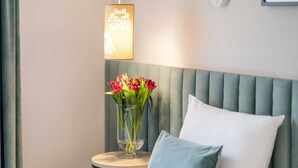 Una cama bien hecha con sábanas blancas, un cabecero tapizado en gris y una mesita de noche con un florero de flores rojas.