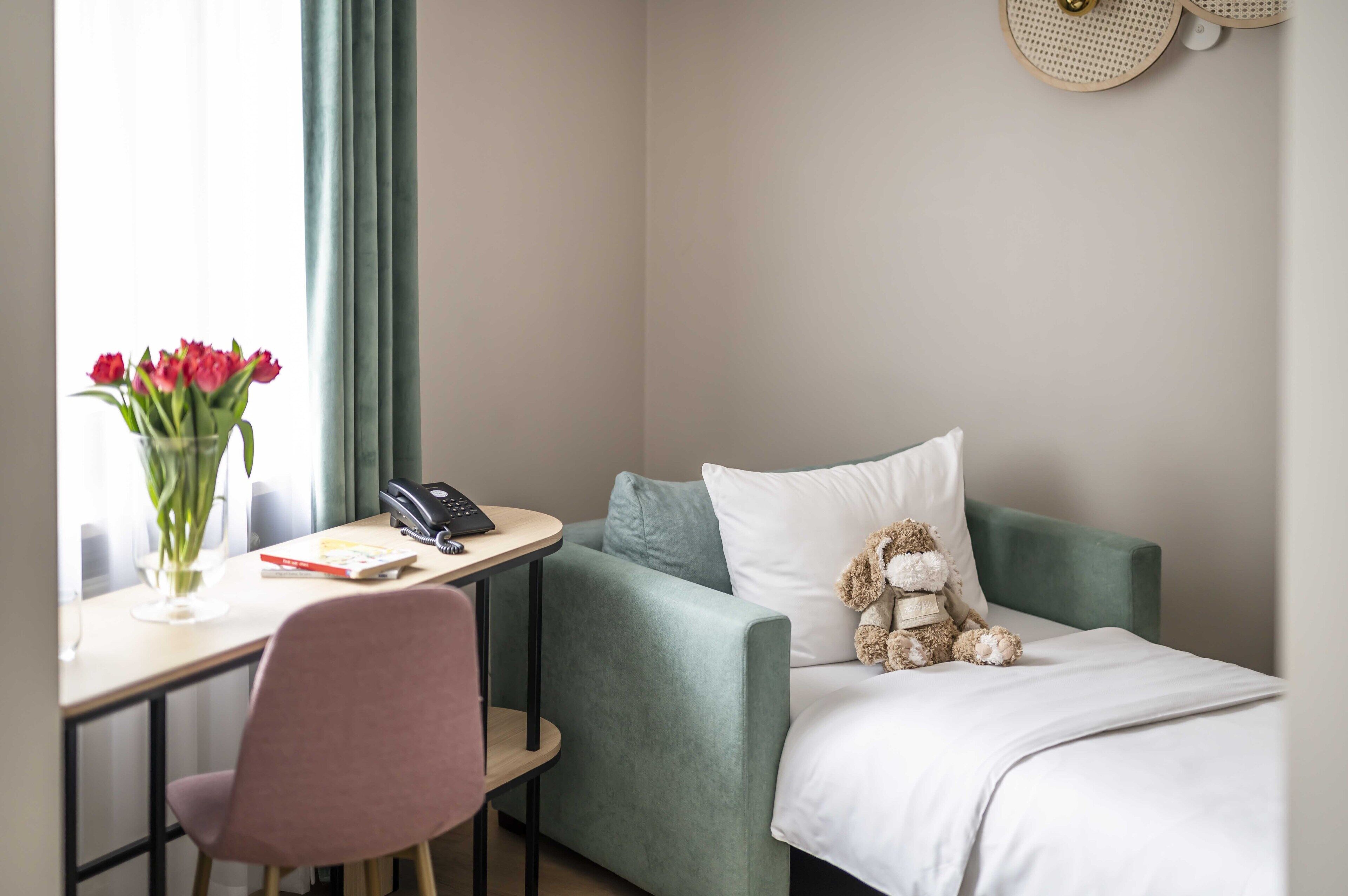 Una habitación de hotel con una cama, un escritorio con una silla, un jarrón de flores y un osito de peluche sobre la cama.
