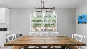 A dining area with a wooden table, white chairs, a hanging light fixture, and a window with blinds. - Dolphin Dreamer-Golf Cart, Pool, BEACH! (Indian Rocks Beach)