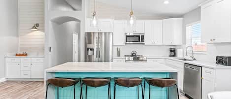 Una cocina moderna con gabinetes blancos, electrodomésticos de acero inoxidable, una isla grande con taburetes de bar e iluminación colgante.