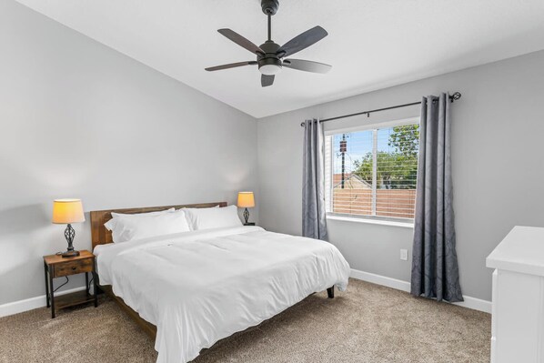 Un dormitorio con cama, dos lámparas de noche, ventilador de techo, ventana con cortinas y cómoda blanca.