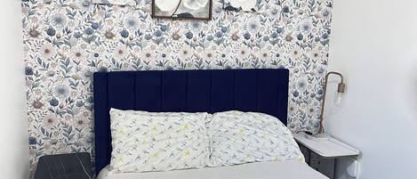 Une chambre à coucher avec un papier peint à motifs floraux, une œuvre d’art encadrée au-dessus du lit, une tête de lit bleu foncé et une table de chevet avec une lampe.