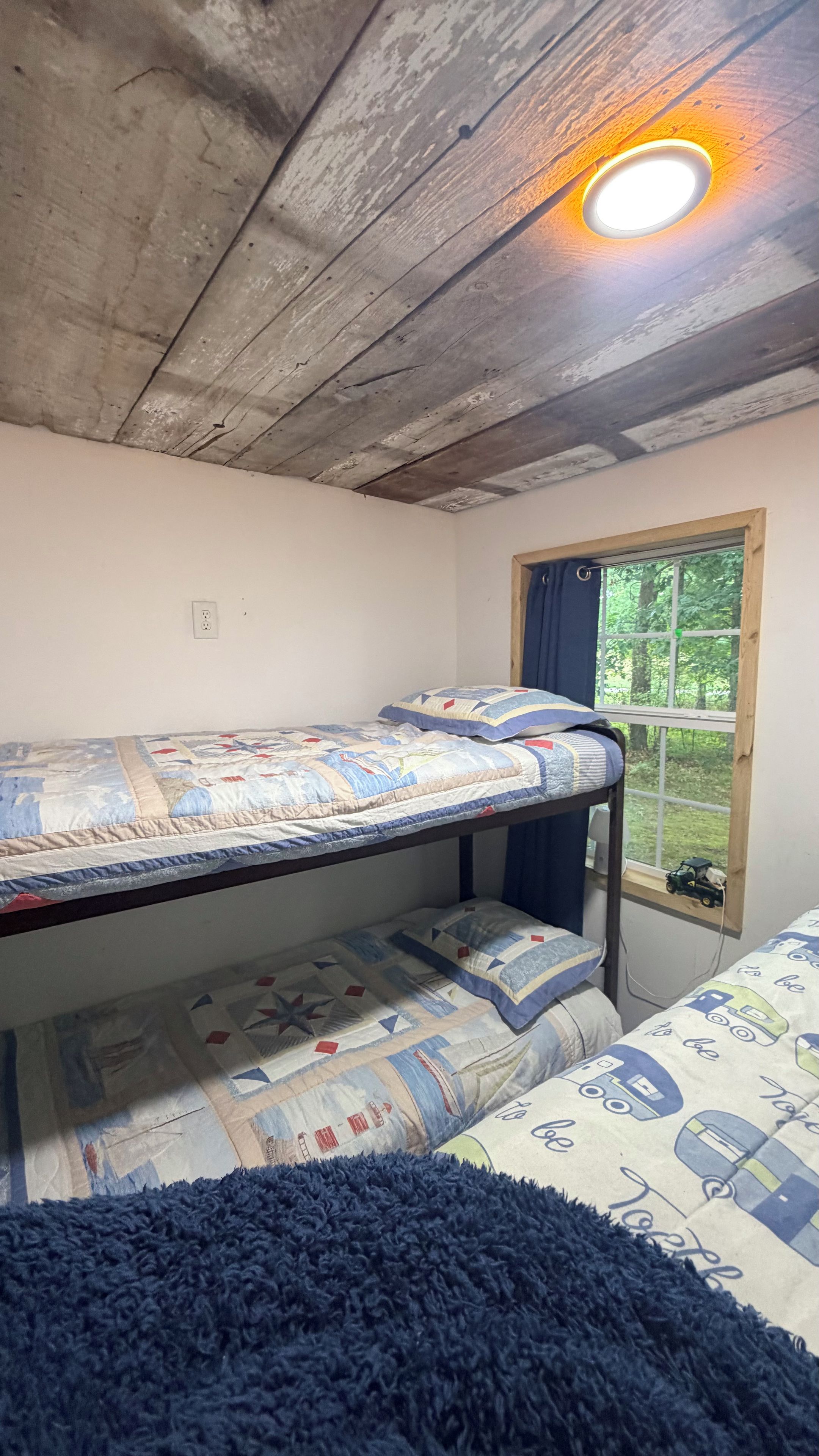 A bunk bed with a patterned quilt, a window with a view of greenery, and a ceiling light.