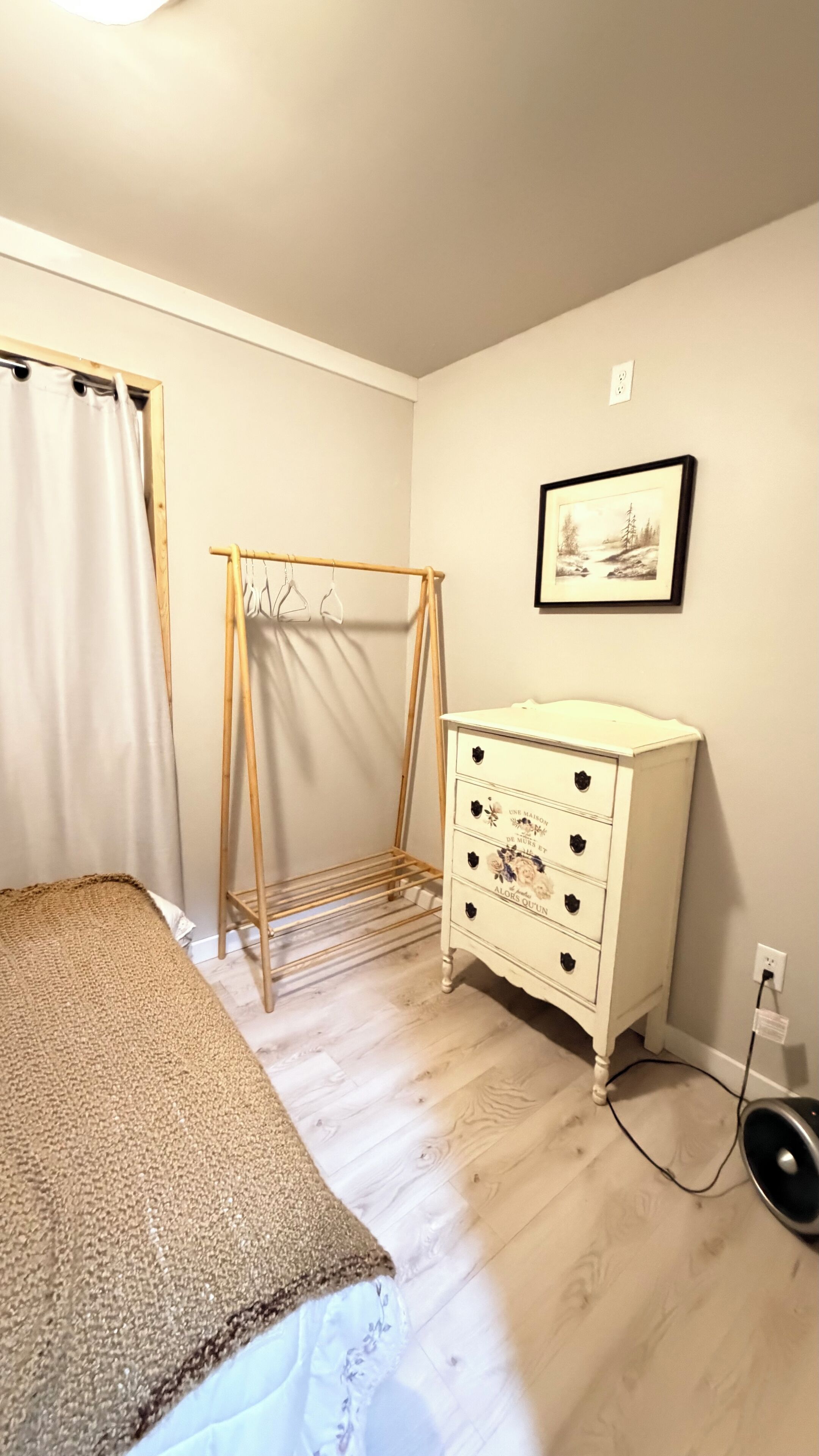 Une chambre avec un lit, un étendoir en bois, une commode blanche et un tableau encadré au mur.