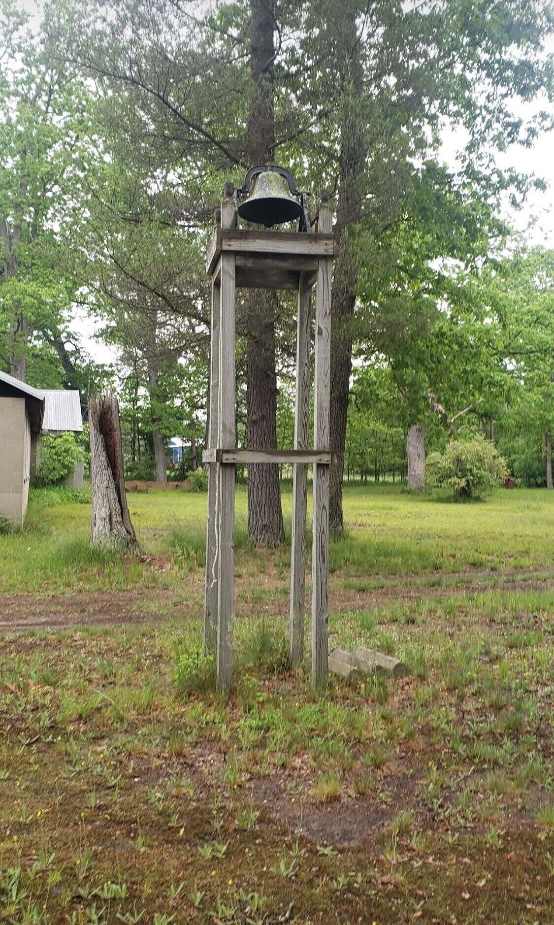 Eine rustikale Holzkonstruktion mit einer Glocke, umgeben von Bäumen und Gras.
