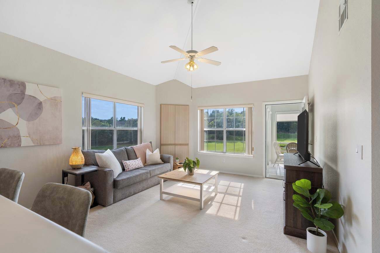 A spacious living room with a ceiling fan, large windows, a sofa, a coffee table, and a TV.