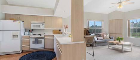 A modern kitchen with white appliances, a dining area with a sofa and coffee table, and large windows offering a view.