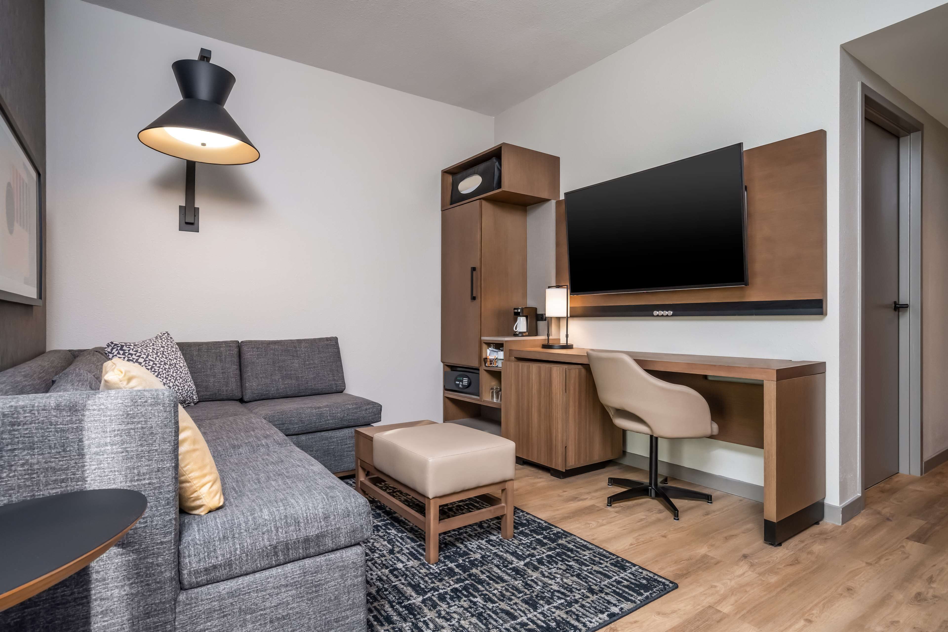 Un salon moderne avec un canapé gris, un petit pouf, un bureau avec une chaise, un téléviseur fixé au mur et une lampe murale.