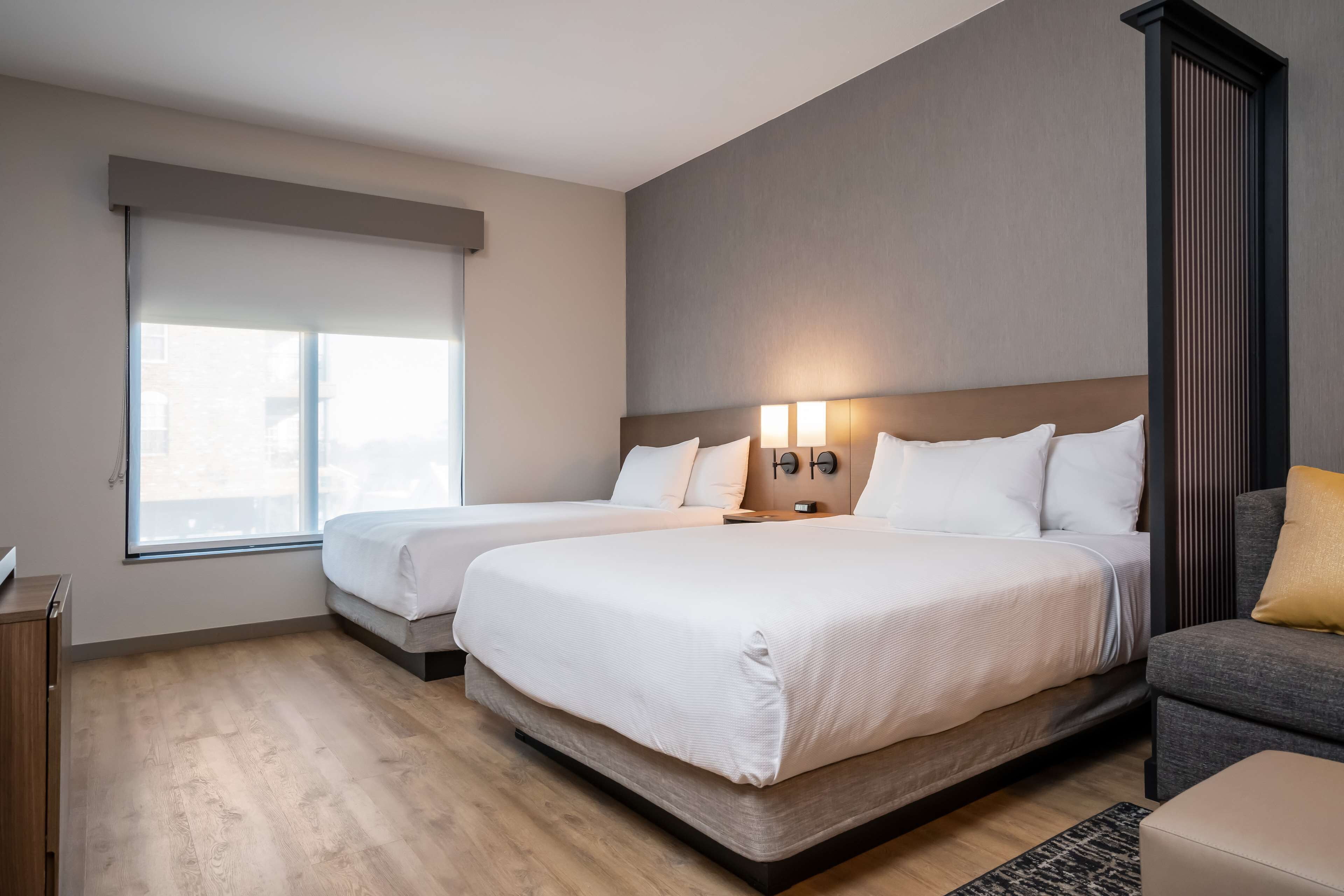 A hotel room with two beds, a sofa, a window with blinds, and a wall-mounted light fixture.