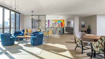 Un hall d’hôtel moderne avec de grandes fenêtres, des sièges bleus et beiges, une fresque murale abstraite aux couleurs vives et une table basse en bois.