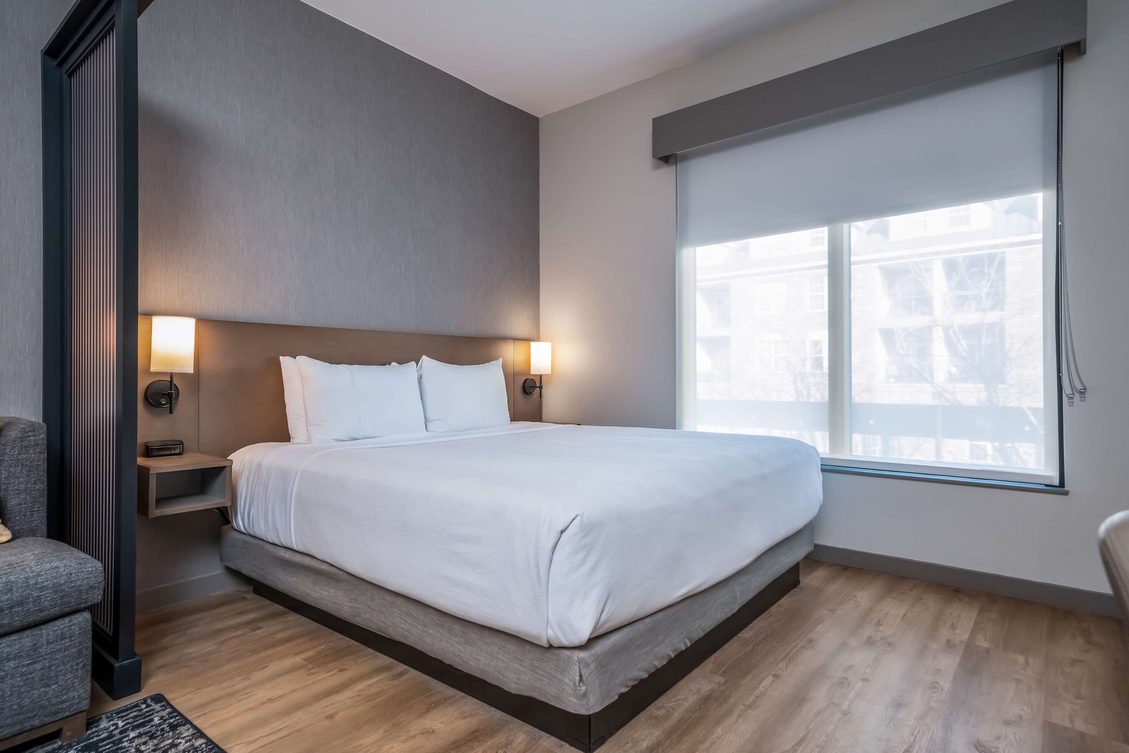 A modern hotel room with a large bed, bedside tables, a chair, and a window with blinds.