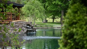A tranquil pond with a stone waterfall, surrounded by lush greenery and a wooden gazebo.