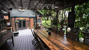 An outdoor wooden deck with a long table and chairs, surrounded by greenery and a building with a sloped roof.